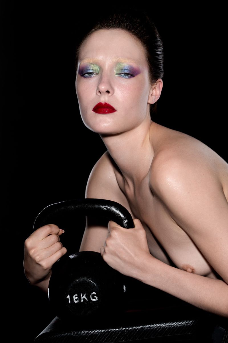 Adult woman in studio with vibrant rainbow makeup, holding black kettlebell on a black textured surface against black backdrop.