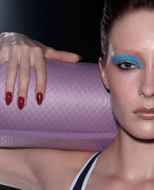 Close-up of a woman with blue eyeshadow and red nails holding a purple yoga mat on her shoulder against a dark background.