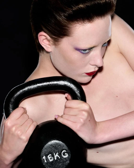 Woman with short brown hair holding a 16kg kettlebell in a studio setting, featuring purple eye makeup and red lipstick against a black background.