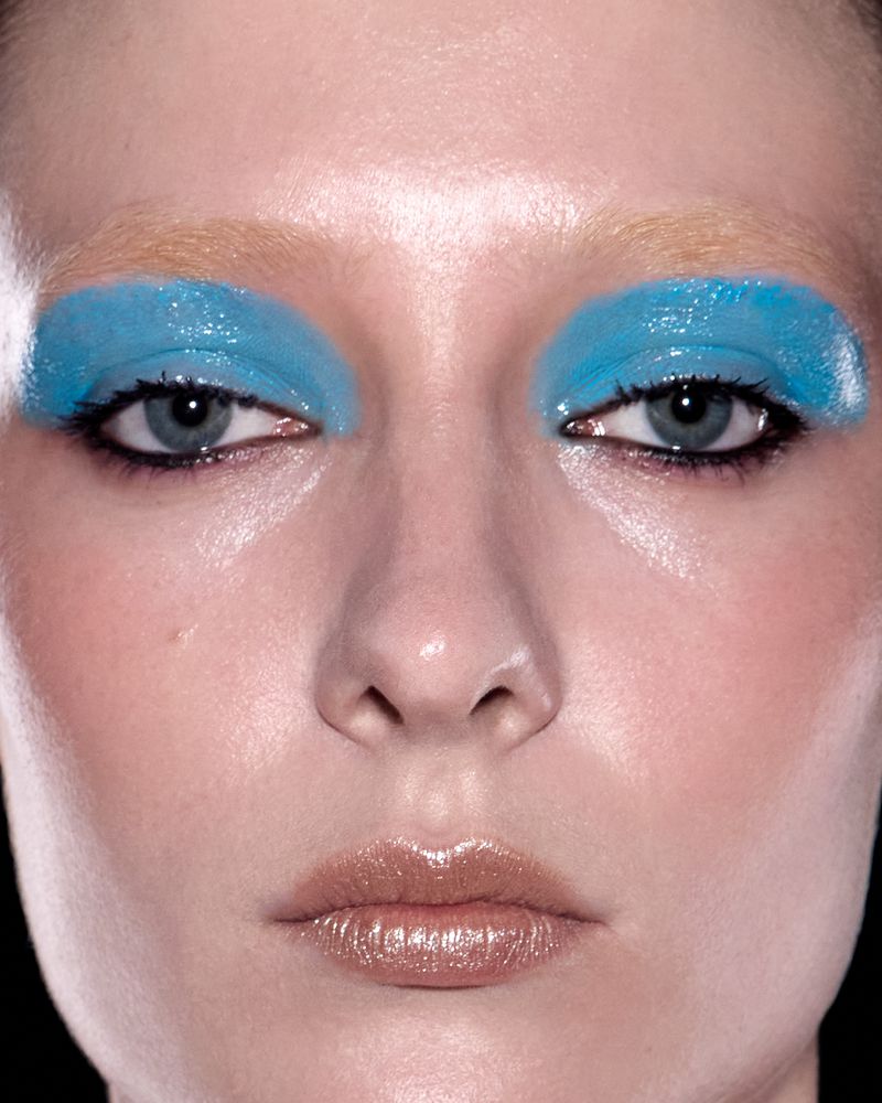 Close-up portrait of a woman with bold blue eyeshadow and glossy lips against a black background, showcasing beauty and makeup.