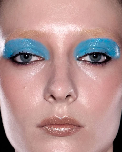 Close-up portrait of a woman with bold blue eyeshadow and glossy lips against a black background, showcasing beauty and makeup.