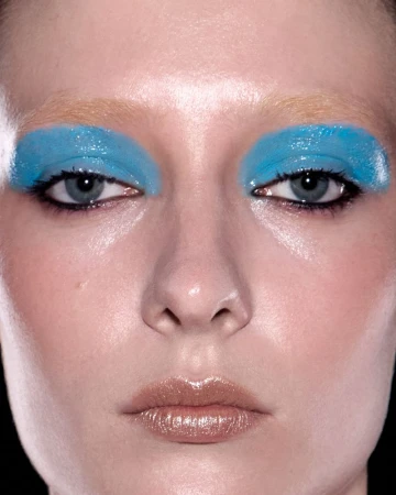 Close-up portrait of a woman with bold blue eyeshadow and glossy lips against a black background, showcasing beauty and makeup.