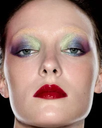 Close-up beauty portrait of a model with vibrant multi-colored eye shadow and glossy red lipstick against a black background.