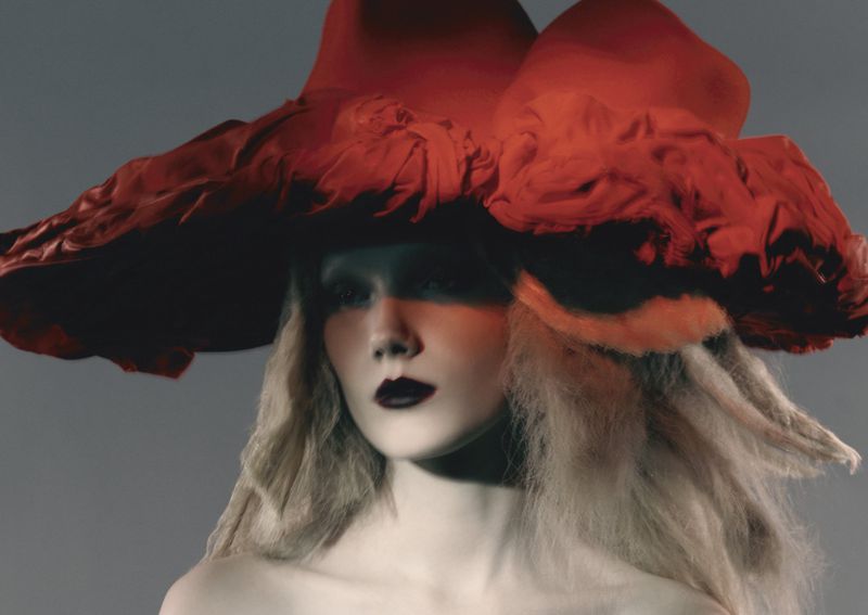 Fashion portrait of a woman with pale skin and light hair wearing a large red hat with black lipstick against a grey background.
