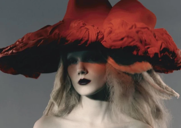 Fashion portrait of a woman with pale skin and light hair wearing a large red hat with black lipstick against a grey background.