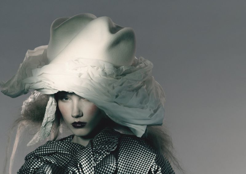 A woman with dark lipstick poses wearing a large sculptural white hat and a black and white checkered jacket in a studio setting.