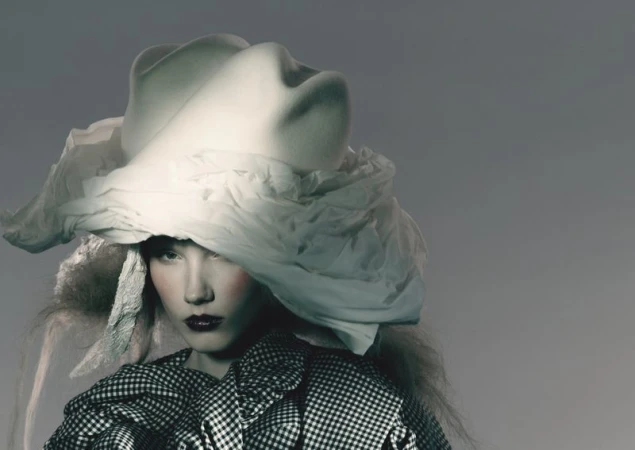 A woman with dark lipstick poses wearing a large sculptural white hat and a black and white checkered jacket in a studio setting.
