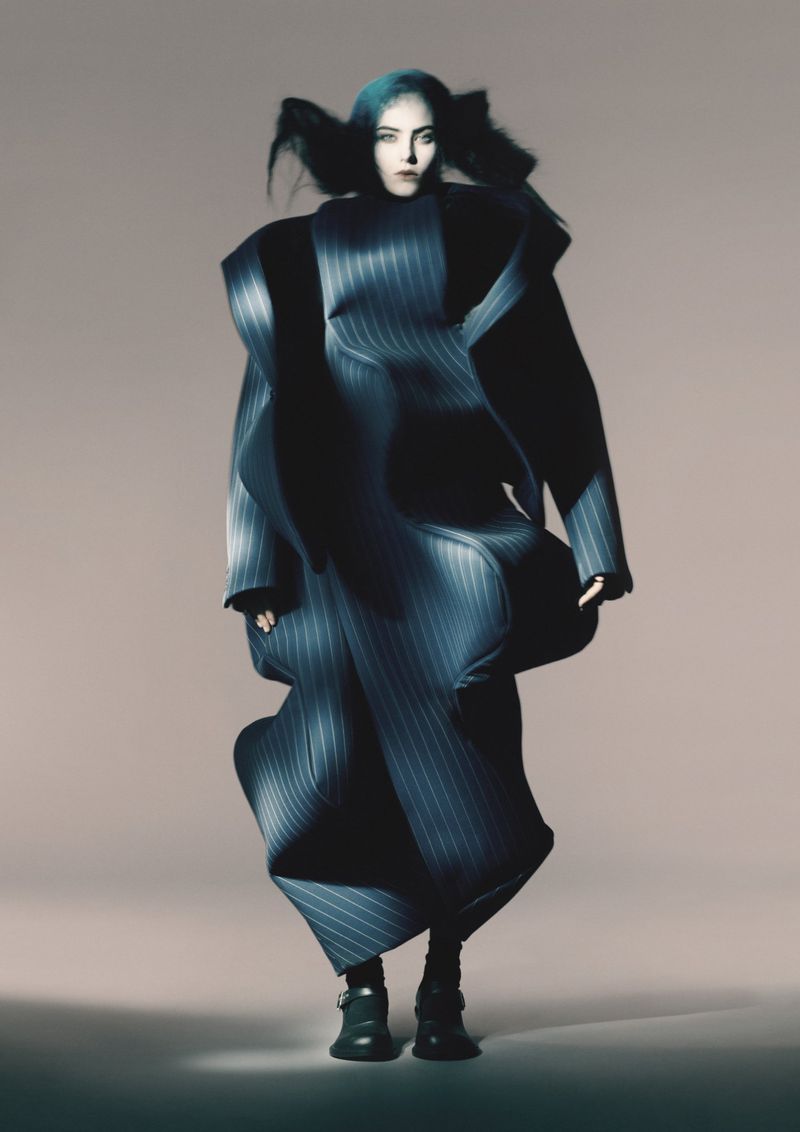 A model wearing a large, pinstripe coat-dress stands in a photo studio with neutral backdrop, showcasing fashion and editorial photography.
