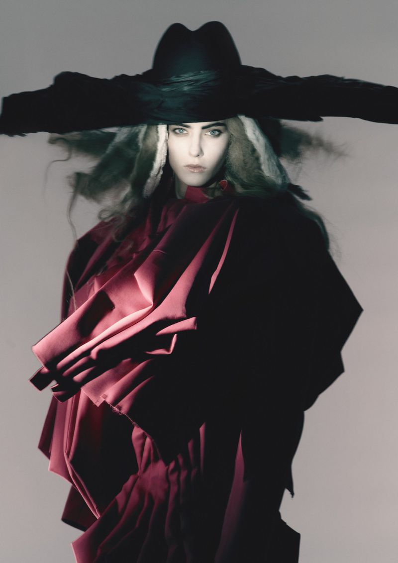 Fashion portrait of an adult female wearing a wide-brimmed black hat and a voluminous magenta garment against a neutral backdrop.