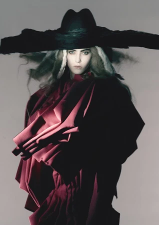 Fashion portrait of an adult female wearing a wide-brimmed black hat and a voluminous magenta garment against a neutral backdrop.