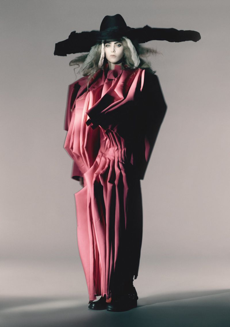 Fashionable adult female poses in a dramatic pink sculptural outfit with oversized black hat in a studio setting. The image has an editorial style.