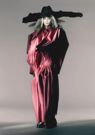 Fashionable adult female poses in a dramatic pink sculptural outfit with oversized black hat in a studio setting. The image has an editorial style.
