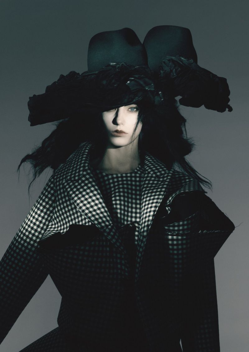 Fashion portrait of a woman wearing a large hat and a houndstooth jacket against a gray backdrop in a studio setting.
