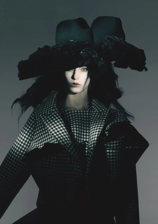 Fashion portrait of a woman wearing a large hat and a houndstooth jacket against a gray backdrop in a studio setting.