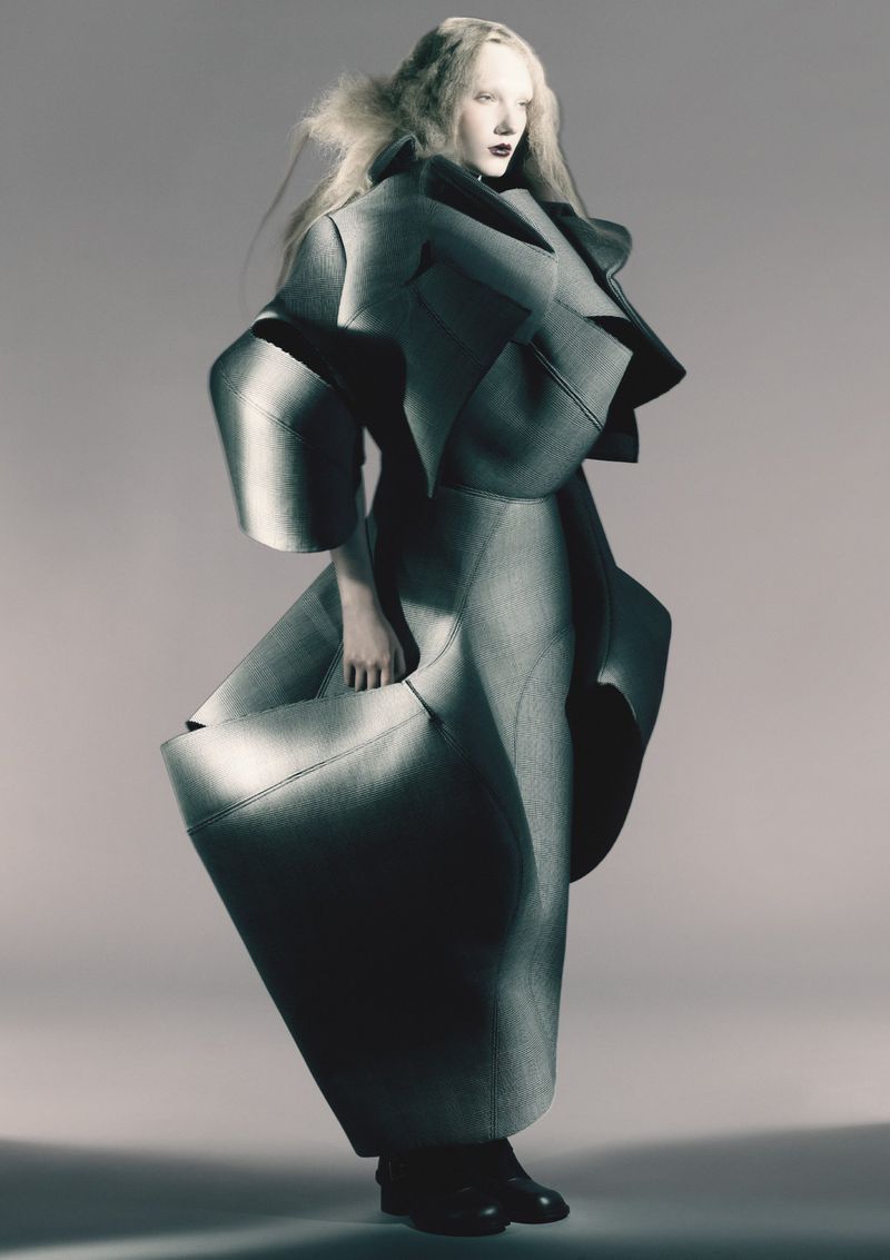 A full-length shot of a fashion model posing in a dramatic grey textured avant-garde coat and skirt with sculptural, angular shapes in a studio.