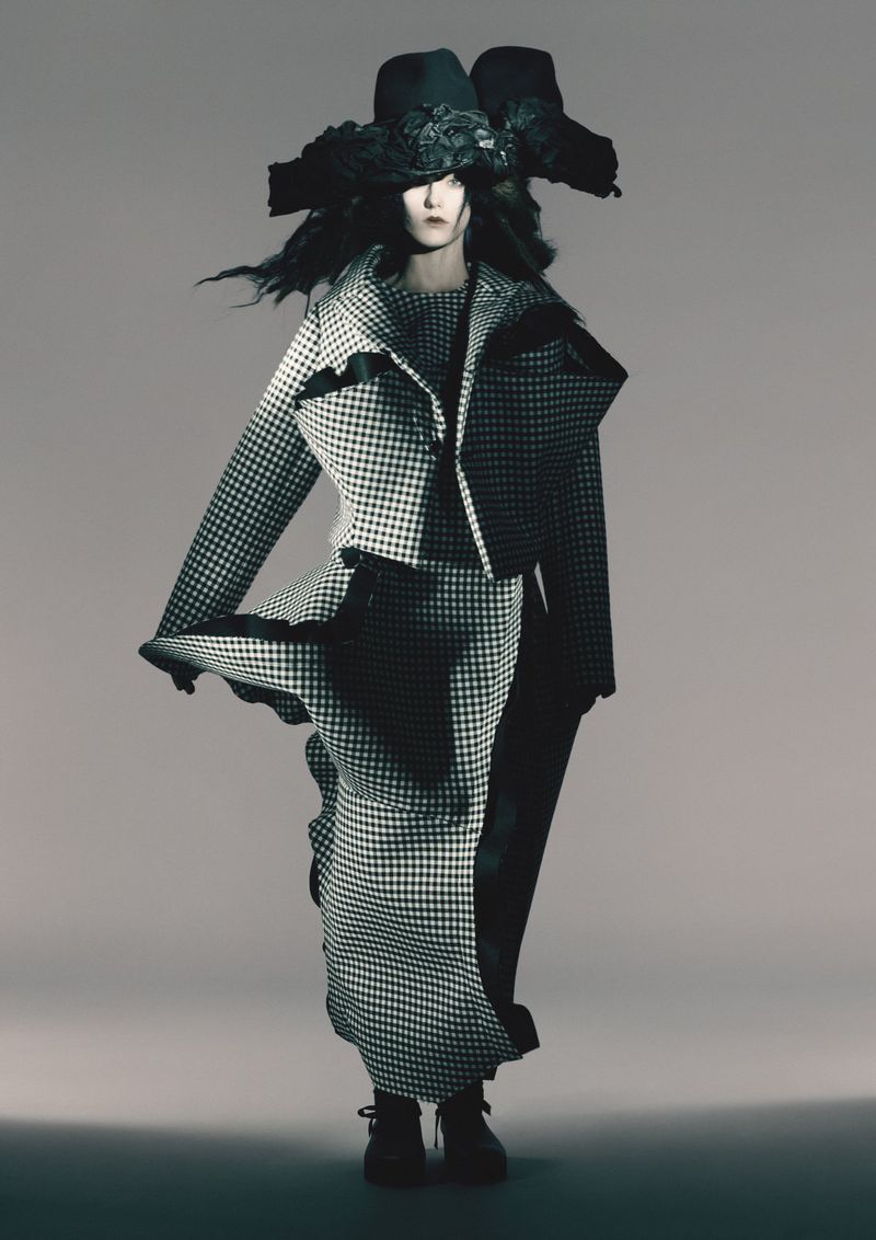 Fashion model in houndstooth jacket and long skirt with dramatic hat, studio shot, monochromatic aesthetic. Runway fashion concept.