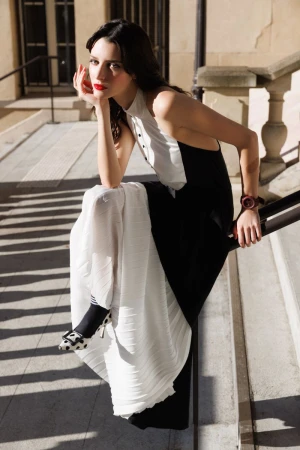 Elegant woman in a black and white dress poses on stone steps with ornate railing. She wears polka dot heels and leans on her hand.