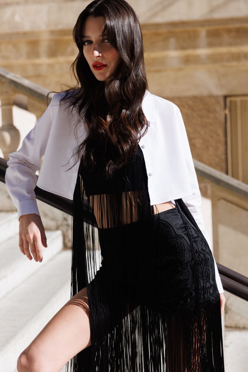 A brunette adult female poses in a black fringe outfit and white jacket on outdoor stairs, leaning on a railing in bright natural light.