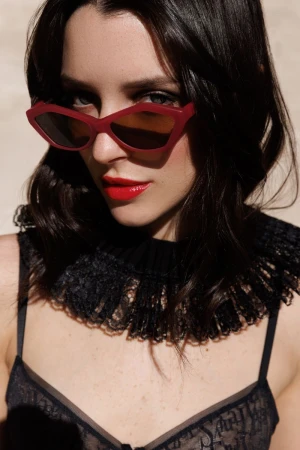 Portrait of a woman with red sunglasses and red lipstick wearing a sheer black top with a lace collar against a neutral background.