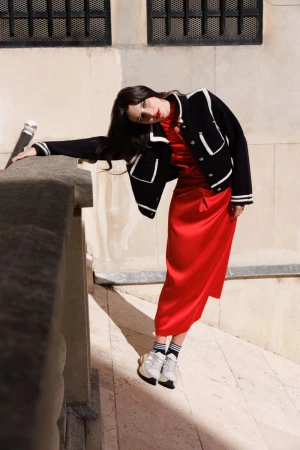 A woman in a red dress and black jacket poses against a stone wall, styled for fashion, with an outdoor urban environment.