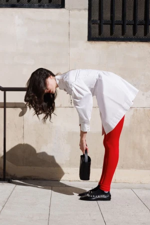 A young adult woman in a white shirt dress and red leggings bends over, holding a black handbag, with a city building in the background.