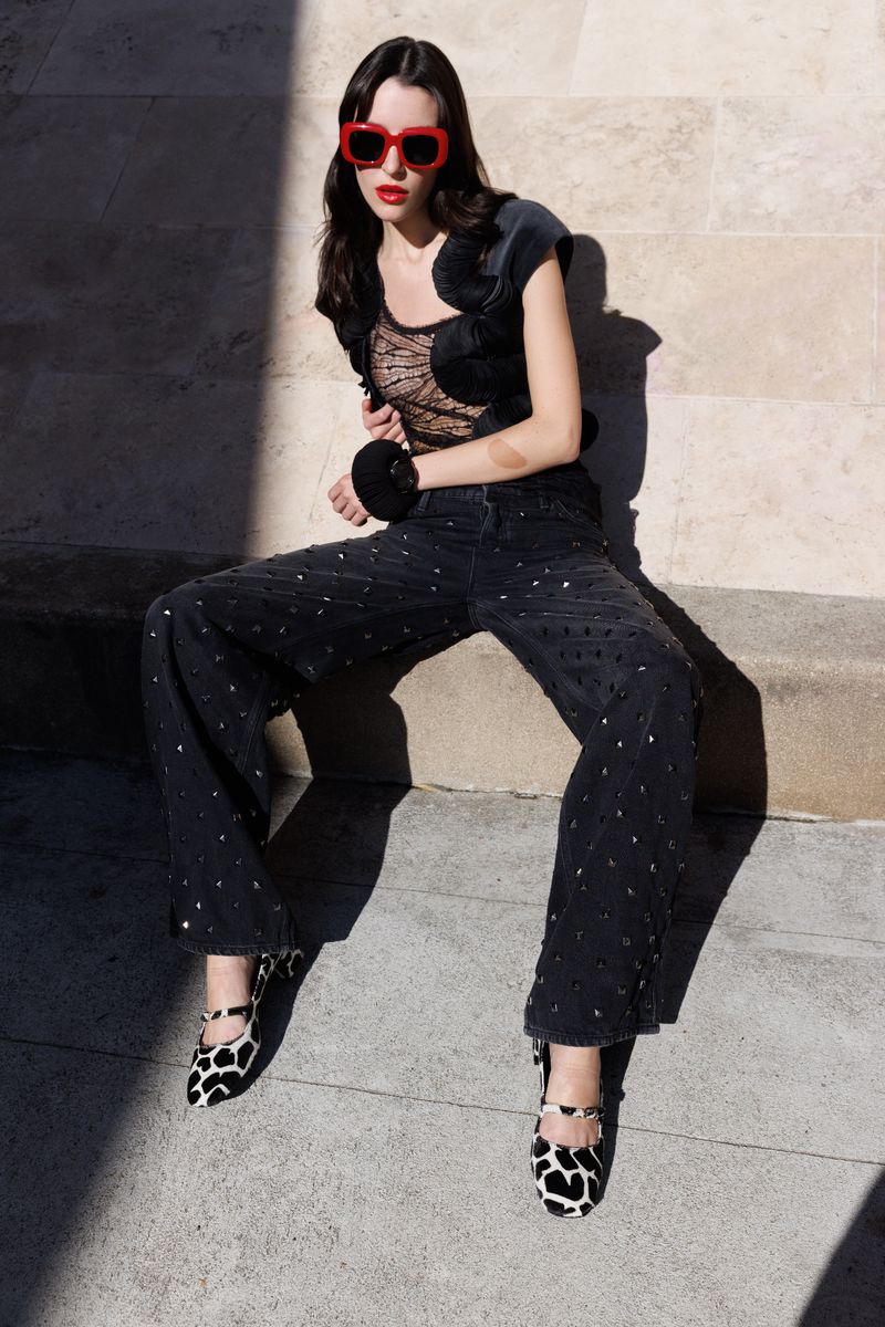 A stylish young woman poses outdoors, wearing trendy red sunglasses, a black lace top, wide-leg jeweled jeans, and black and white cow-print shoes.