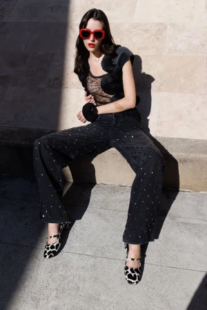 A stylish young woman poses outdoors, wearing trendy red sunglasses, a black lace top, wide-leg jeweled jeans, and black and white cow-print shoes.