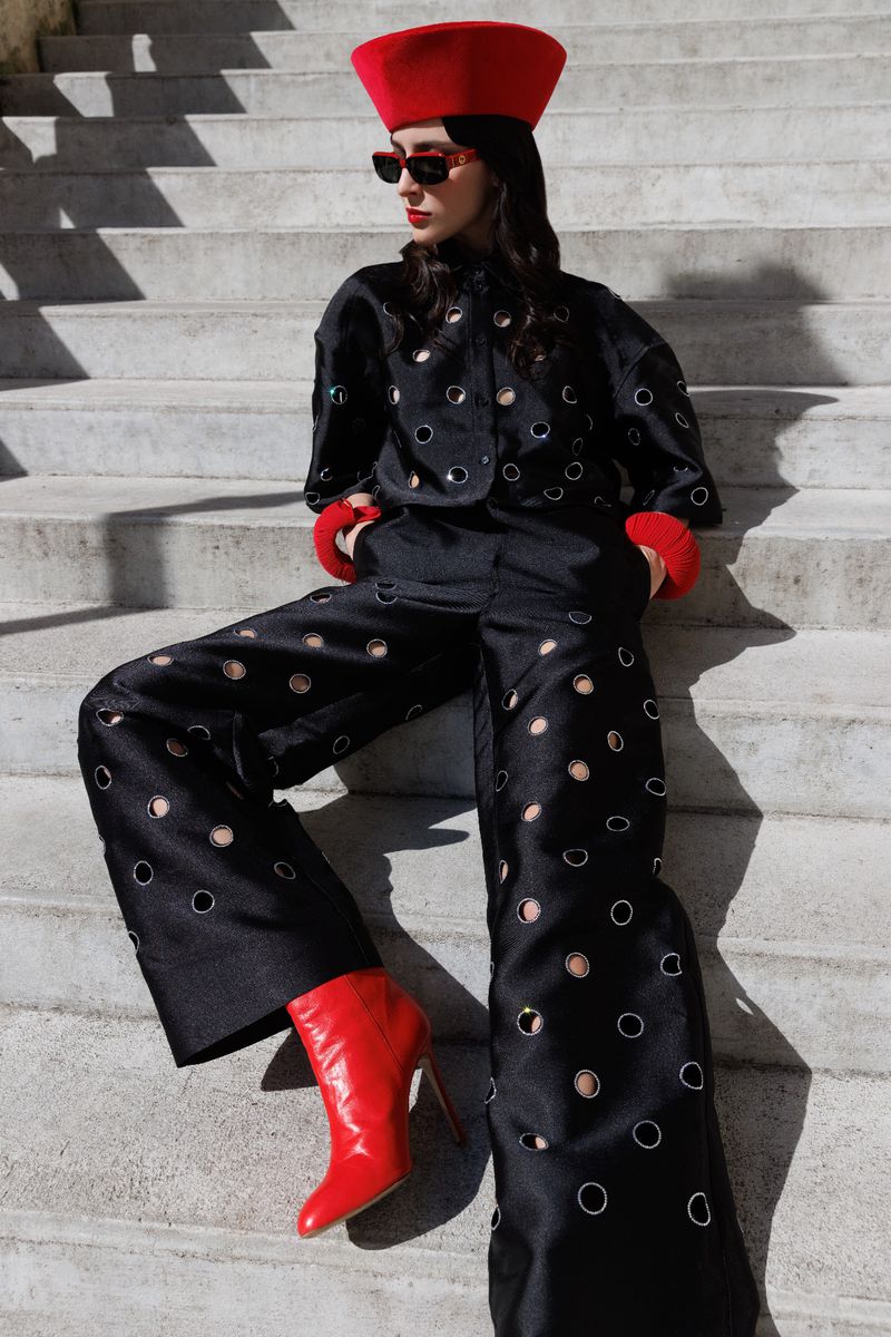 A stylish young woman poses on concrete steps in a black jacket and wide-leg pants with circular cutouts, accessorized with red hat and boots.