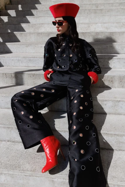 A stylish young woman poses on concrete steps in a black jacket and wide-leg pants with circular cutouts, accessorized with red hat and boots.