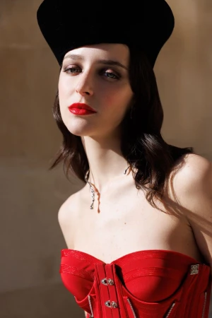 Elegant woman poses in a red corset and black hat, showcasing dark wavy hair, bright red lipstick, and a striking jewelry necklace.