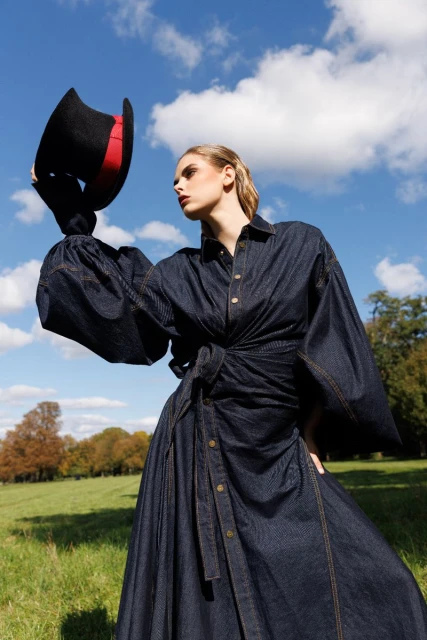 Blonde woman in denim dress and black top hat poses in a field with a blue sky and white clouds. Outdoor fashion shot.