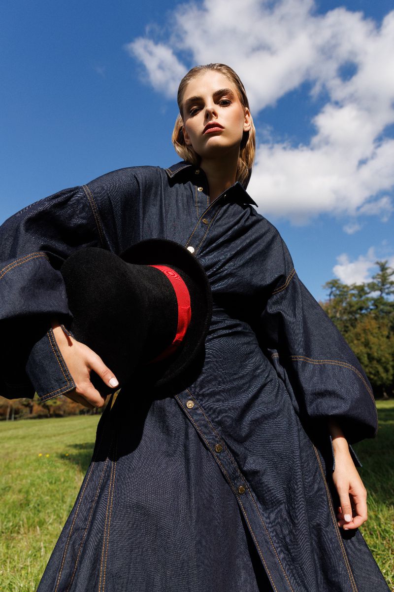 Woman poses in a denim dress with a black hat on a grassy field under a blue, partly cloudy sky, captured in a medium shot.