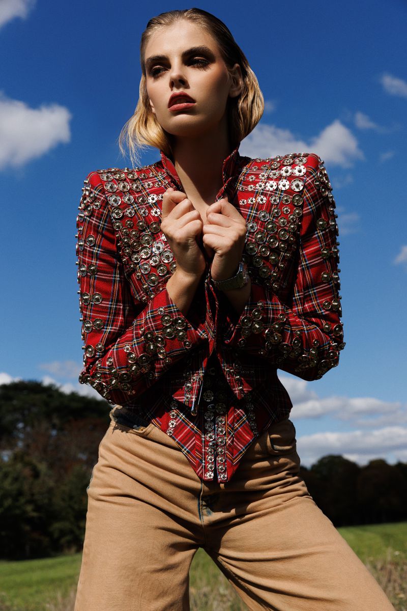 Adult woman in a red tartan jacket with metal hardware, standing outdoors in a field with a blue sky and clouds.