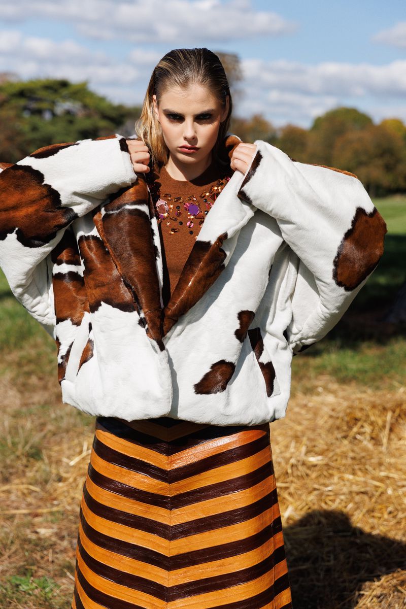 A young woman poses outdoors in a cow-print jacket and striped leather skirt, set against a backdrop of grass and blue sky.