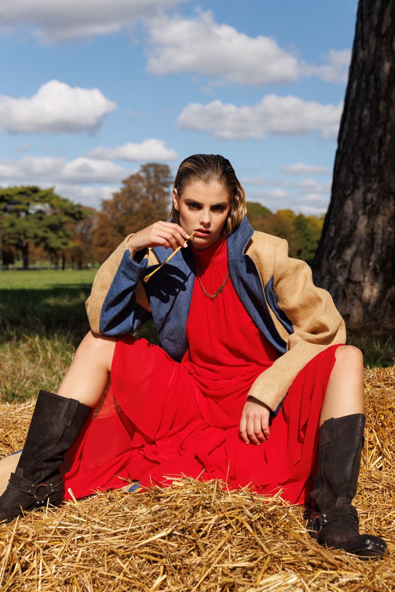 A woman in a red dress and boots sits on hay in a field, posing confidently against a backdrop of blue sky, white clouds, and trees.