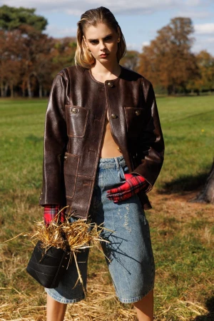 Woman in brown leather jacket and denim shorts holding a small black bucket bag filled with dry grass in a park.