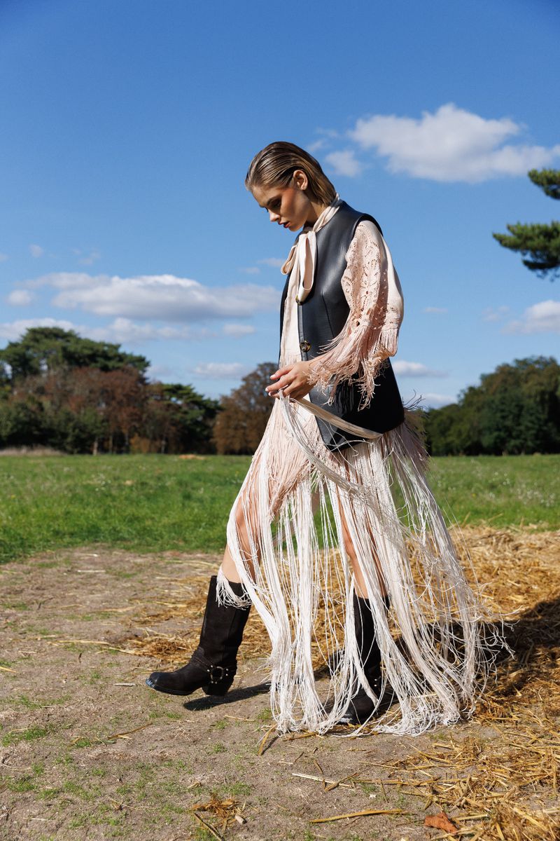 Model in a fringed dress and black vest walking outdoors in a grassy field under a bright blue sky.