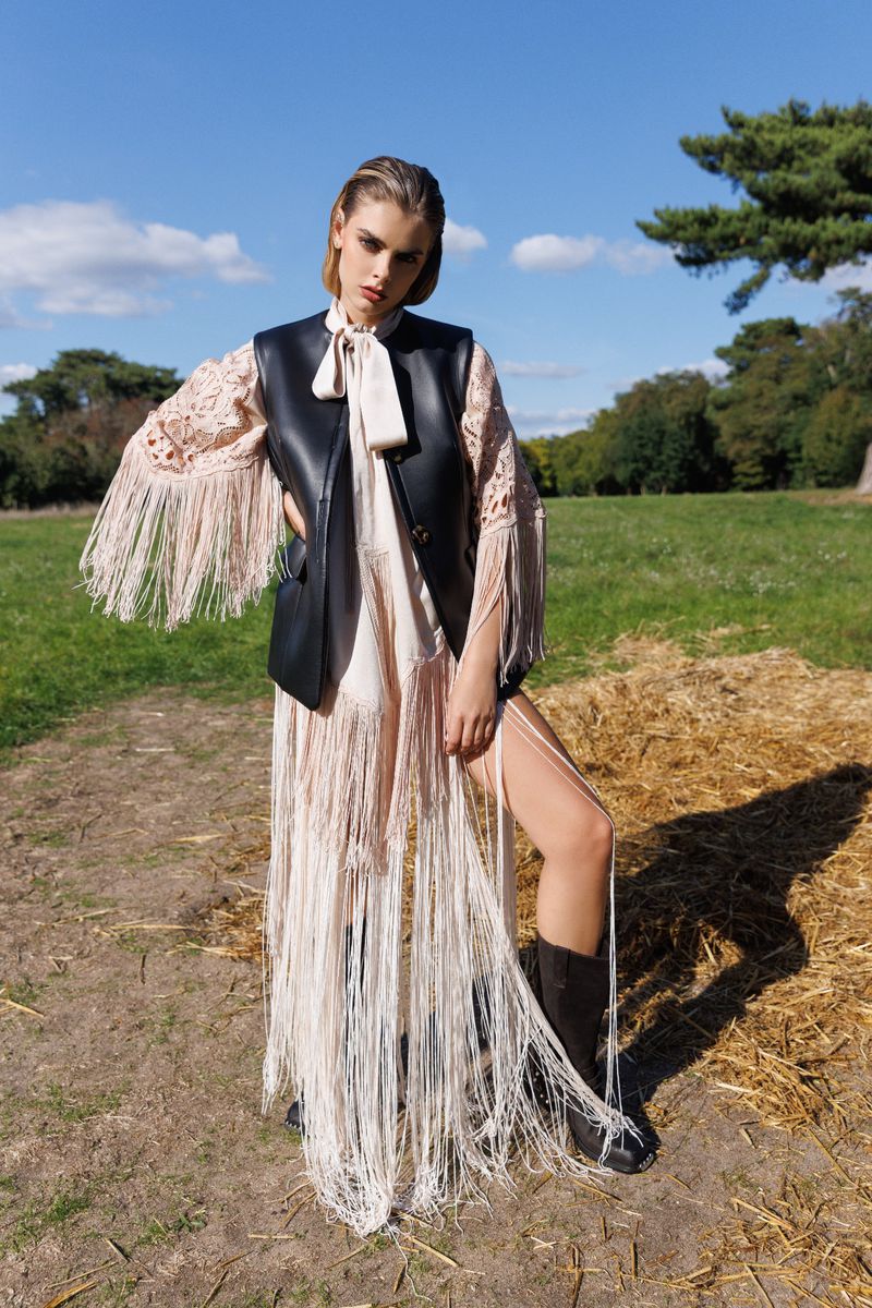 Fashion shot of a woman wearing a leather vest and fringed dress in a field with a blue sky and trees in the background.