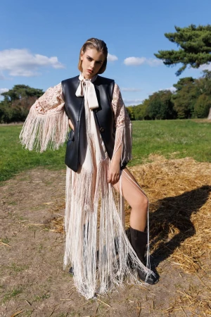 Fashion shot of a woman wearing a leather vest and fringed dress in a field with a blue sky and trees in the background.