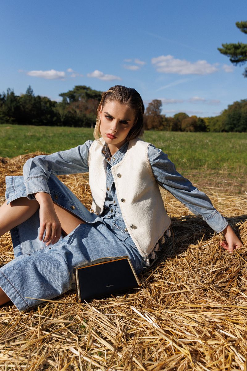 A woman poses sitting on hay wearing a denim dress and a white vest with a black clutch in a green field on a sunny day.