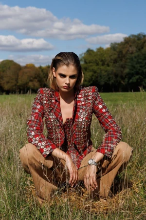 Model in red plaid embellished jacket and brown trousers crouching in a grassy meadow under a cloudy blue sky.