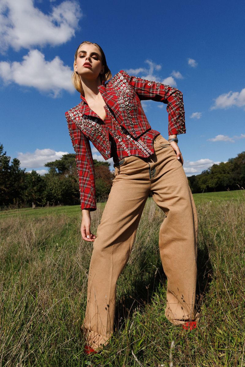 Woman poses in red plaid jacket and tan pants in a grassy field under a blue sky. Fashion editorial shot with natural outdoor lighting.