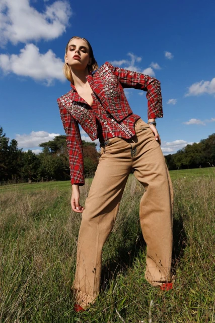 Woman poses in red plaid jacket and tan pants in a grassy field under a blue sky. Fashion editorial shot with natural outdoor lighting.