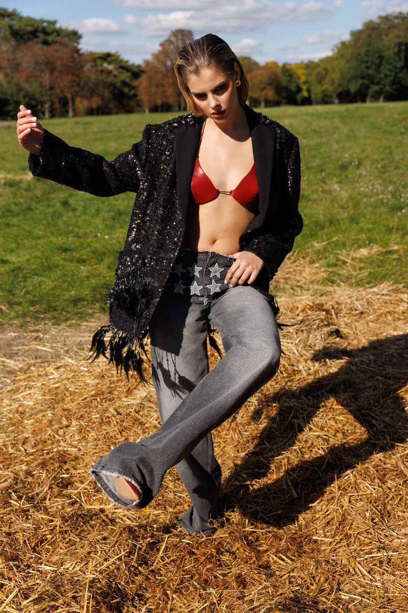 A young woman poses in an open field wearing a sequined jacket, gray pants and a red bra, with trees visible in the background under a sunny sky.