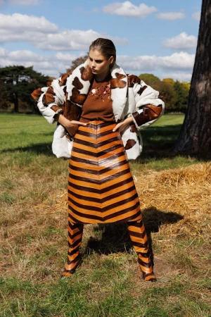 Model standing in a grassy field wearing a cow-print coat, brown knit top, striped skirt, and matching knee-high boots.