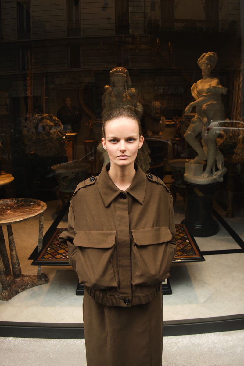 An adult female poses in a knee-length, olive green jacket and skirt in front of a storefront window displaying statuary.