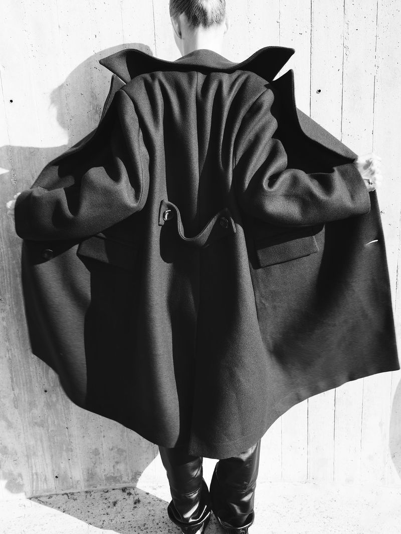 Fashion shot of a person with short hair from behind wearing an oversized black coat with shiny black boots against a concrete wall.