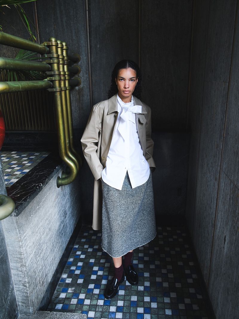 A stylish adult female stands indoors wearing a light beige trench coat, a white collared shirt with a tie, and a gray textured skirt.