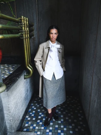 A stylish adult female stands indoors wearing a light beige trench coat, a white collared shirt with a tie, and a gray textured skirt.