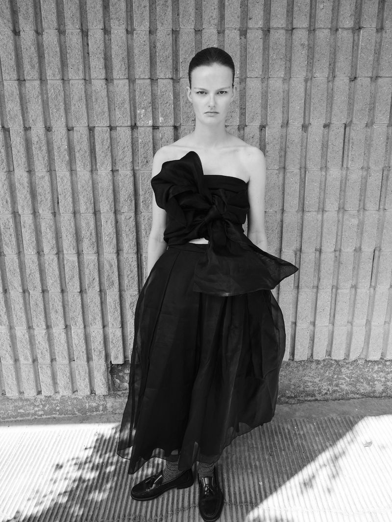 Monochrome fashion portrait of a model wearing a black strapless dress with a large bow detail against a textured brick wall.